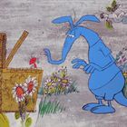 Eine blaue cartoonhafte Ameise betrachtet interessiert ein Picknickkorb neben bunten Blumen im Gras.