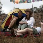 Drei Personen sitzen und liegen entspannt auf einer Decke unter einem bunten Sonnenschirm in einer trockenen, kargen Landschaft.