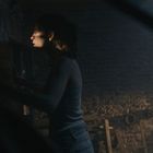 Keine Texte sichtbar. Eine Frau in einem dunklen Raum, schwaches Licht auf ihr Gesicht, Steinmauer im Hintergrund.