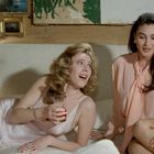 Zwei Frauen sitzen lächelnd auf einem Sofa, eine in einem Nachthemd mit einem Getränk, die andere in einer Bluse.