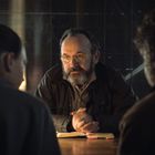 Ein Mann mit Brille und Bart sitzt an einem Tisch, deutet nachdenklich. Zwei Personen sind ihm zugewandt.