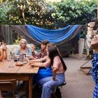 Mehrere Frauen sitzen um einen Tisch, eine Frau steht rechts, hält eine Zigarette. Hintergrund: grüne Pflanzen.