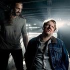 Zwei Männer in einem dunklen Raum: Einer mit bedrücktem Ausdruck sitzt, der andere steht bedrohlich daneben. Lichtstrahlen im Hintergrund.