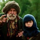 Ein älterer Mann mit Wollmütze zeigt, steht mit einem Kind in blauer Mütze im Freien. Dichte grüne Bäume im Hintergrund.