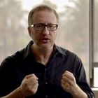 Ein Mann mit Brille und Bart trägt ein dunkles Hemd und hält die Fäuste erhoben. Im Hintergrund ist ein Fenster.