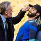 Zwei Männer stehen nahe beieinander. Der linke Mann stützt seinen rechten Arm gegen die Stirn des rechten Mannes, der eine Kappe trägt.