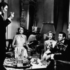 Eine elegante Gesellschaftsszene in einem luxuriösen Raum: Fünf Menschen in Abendgarderobe mit Cocktails in festlicher Stimmung.
