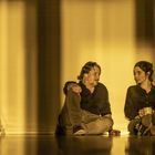 Zwei Personen sitzen am Boden, eine Leiter links, gelbliche Beleuchtung. Eine hält eine Tasse, daneben steht eine Flasche.