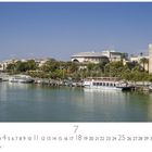 Aufschrift: "Sevilla". Szene: Fluss mit Booten, grüne Uferpromenade und ikonischer Turm unter blauem Himmel.