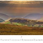 "Galway", Kalenderseite für Juli, Hügellandschaft mit Lichtstrahlen durch Wolken, grüne Felder und Berge im Hintergrund.