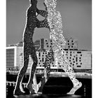 Text unten: "Molecule Man". Kalender mit Tagen und Datum. Skulptur zweier Figuren aus durchlöchertem Metall vor moderner Architektur.