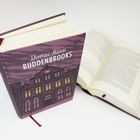 "Thomas Mann, Buddenbrooks" auf einem Buchcover. Gezeichnetes Gebäude mit gelben Fenstern auf violettem Hintergrund.