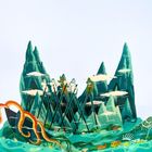 Es ist eine bunte, detaillierte Illustration mit Charakteren auf Stelzen in einer fantasievollen Landschaft vor Bergen und Wasser.