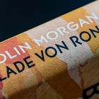 Text: "Colin Morgan Ballade von Ronan". Hintergrund in Rottönen mit einem abstrakten Design.