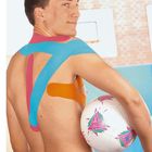 Ein Mann mit buntem Kinesiotape auf dem Rücken hält einen Fußball unter dem Arm in einer Sporthalle.