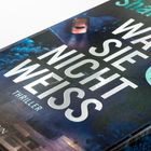 „Was sie nicht weiß“ von Shari Lapena, ein Thriller. Im Hintergrund ist ein Bild einer Straße mit Auto.