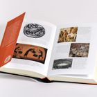 Ein geöffnetes Buch mit Abbildungen und Text über historische Themen. Der Umschlag ist rot mit weiterem Text.