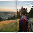 AUSZEIT. ABENTEUER. LEBENSFREUDE. Ein Radfahrer mit Helm blickt über eine weite Landschaft bei Sonnenuntergang.