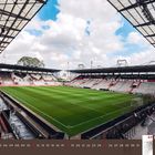 07 2027, Juli. Stadion mit Texten: "Kein Mensch ist illegal", "Kein Fußball den Faschisten". Leere Tribünen, grüner Rasen.