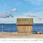 Kalender für Juli. Holzhütte am steinigen Ufer mit blauen Meer und Himmel. Kleine Boote am rechten Bildrand.