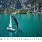 Unter dem Bild ist der Monatskalender für Juli. Ein Segelboot auf türkisfarbenem See, umgeben von bewaldeten Bergen.