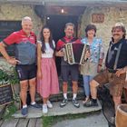 Eine Gruppe von fünf Menschen steht vor einer Hütte. Einer hält ein Akkordeon. Holzschilder zeigen Speisen wie Frittatensuppe.