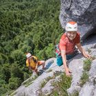 Eine ältere Frau klettert an einer Felswand, mit Helm und Kletterausrüstung. Unten im Bild ein weiterer Kletterer.