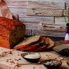 Ein ungeschnittenes Brot mit einigen geschnittenen Scheiben. Daneben Schalen mit Samen, Körnern und Dekovasen mit Blumen.