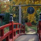 Rote Holzbrücke führt zu einem Garten mit herbstlichen Bäumen und einem Gong, umrahmt von üppigem Grün.