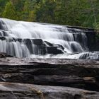 Ein breiter Wasserfall fließt über gestufte Felsen, umgeben von üppigem Grün.