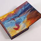 Gemälde in einem geöffneten Buch: Eine Person steht am Meer, umgeben von Bergen, fliegenden Kreaturen und einem farbenfrohen Himmel.