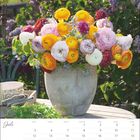 Kalenderseite Juli. Ein Blumenstrauß mit bunten Ranunkeln steht auf einem Tisch im Freien, umgeben von lila Blüten.