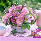 "Juli" ist groß geschrieben. Ein Blumenarrangement in Rosa mit Tasse und Geschenk auf gedecktem Tisch. Kalenderdaten sichtbar.