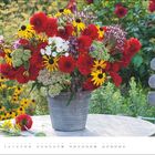 JULI, 31 Tage. Ein bunter Blumenstrauß in einem Eimer steht auf einem Tisch im Garten, mit roten und gelben Blumen.