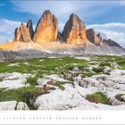 „JULI“ am unteren Rand. Ansicht von Berggipfeln, Wiese mit Steinen im Vordergrund, blauer Himmel mit Wolken.
