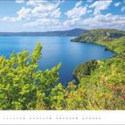 JULI, darunter Monatskalender. Landschaft mit blauem See, grüne Hügeln im Hintergrund, vorne blühende Sträucher.
