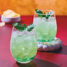 Zwei Gläser mit grünem Getränk, Eiswürfeln und Basilikum auf rotem Tisch; unscharfer Hintergrund mit Snackschale.