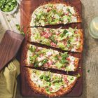 Ein rechteckiger Flammkuchen auf Holzbrett, mit Schinken, Lauchzwiebeln und Kräutern garniert, daneben ein Glas Wasser.