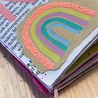 Ein buntes, handgemachtes Notizheft mit Zeitungsausschnitten, rosa Stoff und einer Regenbogen-Collage auf grauem Karton.