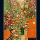 Eine Frau steht vor einem blumigen Hintergrund, bekleidet mit einem bunten, gemusterten Gewand. Kalender unten.