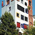 Kalenderseite mit Juli 2027; Zahlen in Weiß und Orange. Ein farbenfrohes Gebäude im Hundertwasser-Stil mit vielen Bäumen.