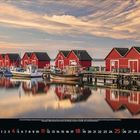 Kalenderseite mit Juli-Tagen. Rote Holzhäuser und Boote spiegeln sich im Wasser. Harmonische Abendstimmung.