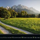 "7 2024" und Kalendertage. Ein Weg führt durch eine grüne Wiese, dahinter Bäume und ein Gebirge bei klarem Himmel.