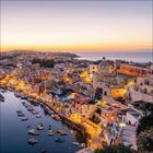 Keine Texte vorhanden. Abenddämmerung über einer malerischen Küstenstadt mit bunt beleuchteten Häusern und Booten im Wasser.