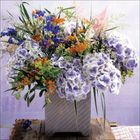 Text: Keiner sichtbar. Ein üppiger Blumenstrauß in Pastelltönen, arrangiert in einer eckigen Vase auf einem rustikalen Tisch.