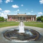 Ein großer Brunnen vor einem historischen Gebäude mit Säulen, flankiert von Grasflächen und blauem Himmel mit Wolken.