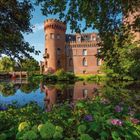 Ein Schloss mit einem runden Turm, umgeben von einem Wassergraben, Bäumen und bunten Blumen im Vordergrund.