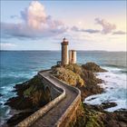 Ein Pfad führt zu einem Leuchtturm auf einer felsigen Landzunge, umgeben vom Meer bei Sonnenuntergang.