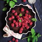 Keine erkennbaren Texte. Ein Schokoladenkuchen mit Himbeeren und Minzblättern auf einem Tuch, ein Messer liegt daneben.