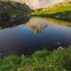 07; eine Bergspitze spiegelt sich in einem ruhigen See, umgeben von grüner Wiese und rosa Blumen.